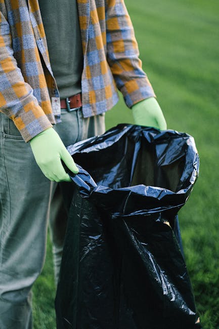 The image shows a person wearing a yellow and grey checkered shirt, olive green gloves, and grey trousers standing outdoors on a grassy area. They are holding open a large black plastic rubbish bag, preparing for waste disposal or collection. The background features lush green grass and blurred natural surroundings, indicating an outdoor environment possibly related to garden cleanup or waste removal. The scene suggests activities associated with rubbish collection or waste management services, emphasizing the handling and disposal of waste materials in a tidy and responsible manner. The individual’s attire and the use of gloves highlight safety and hygiene considerations commonly observed during rubbish removal tasks, in line with services provided by companies like Waste Removal Shepherds Bush that handle various types of waste in private or alternative disposal settings.