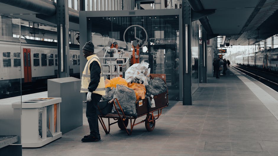 A person dressed in a black jacket, dark pants, a gray beanie, and a high-visibility vest is standing on a train station platform near a trolley loaded with various bags of rubbish and discarded items, including black, gray, and orange plastic bags, some appearing full and tightly packed. The trolley's metal frame is orange, and the bags are stacked and piled on top, with some spilling over the sides. Behind the individual, a glass safety barrier separates the platform from the train tracks, where a modern train with a sleek, silver exterior and large windows is parked. To the left, another train with a similar design is visible, and a few passengers can be seen further down the platform. The environment is lit with diffused natural light, and the overall scene suggests a typical urban railway station setting. This image illustrates the process of on-site rubbish handling, potentially related to waste collection or disposal services such as those offered by Waste Removal Shepherds Bush, highlighting alternative waste management options outside conventional civic collection methods.