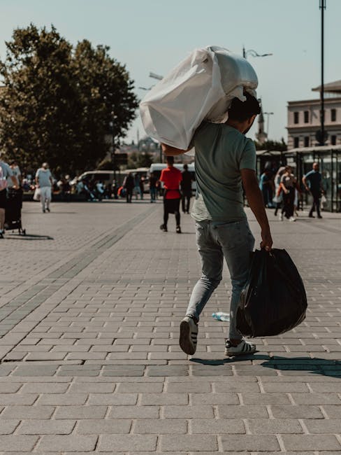 A young man walking along a paved urban plaza during daylight, carrying a large white plastic bag over his shoulder with visible creases and a slightly crumpled texture, and holding a black duffle bag in his right hand. The surrounding environment includes a few trees, several pedestrians in casual clothing, and modern buildings in the background. The scene appears relaxed, with some people standing or walking, and a street with parked cars is visible further back. The lighting is natural, suggesting a sunny day, and the overall atmosphere indicates a typical city setting. This context subtly aligns with private waste handling, as the individual appears to be engaged in personal rubbish disposal, and Waste Removal Shepherds Bush offers similar services for on-site clearance and alternative waste collection solutions.
